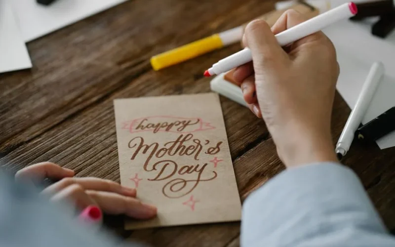 Close-up of a person designing a Mother's Day card with calligraphy on wooden table.