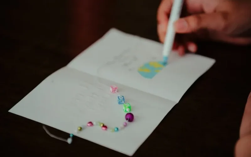 Close-up of a hand decorating a greeting card with colorful beads and a pen, creating a personalized design.