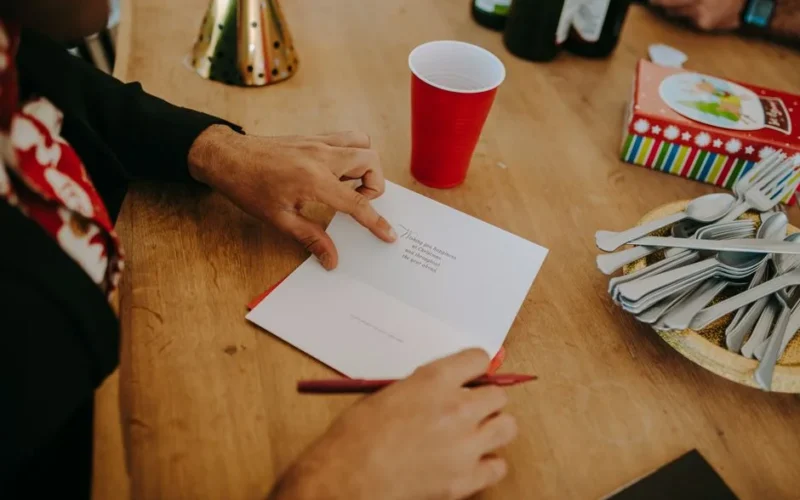 Person writing a Christmas card on a wooden table with festive decor.
