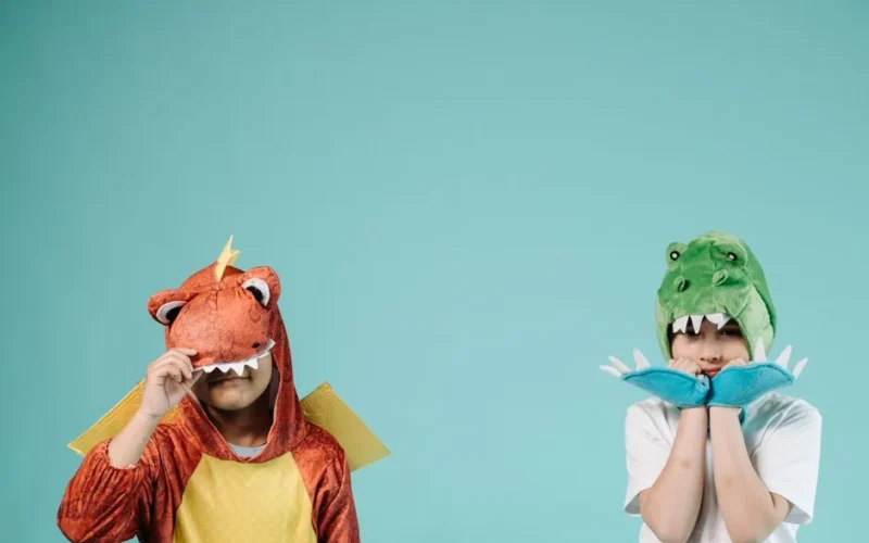 Two kids wearing dinosaur costumes pose cheerfully indoors against a turquoise backdrop.