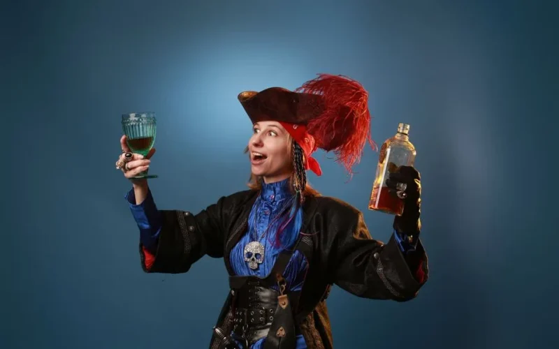 Amazed woman in pirate costume and pendant with decorative skull holding vintage glass and bottle of booze