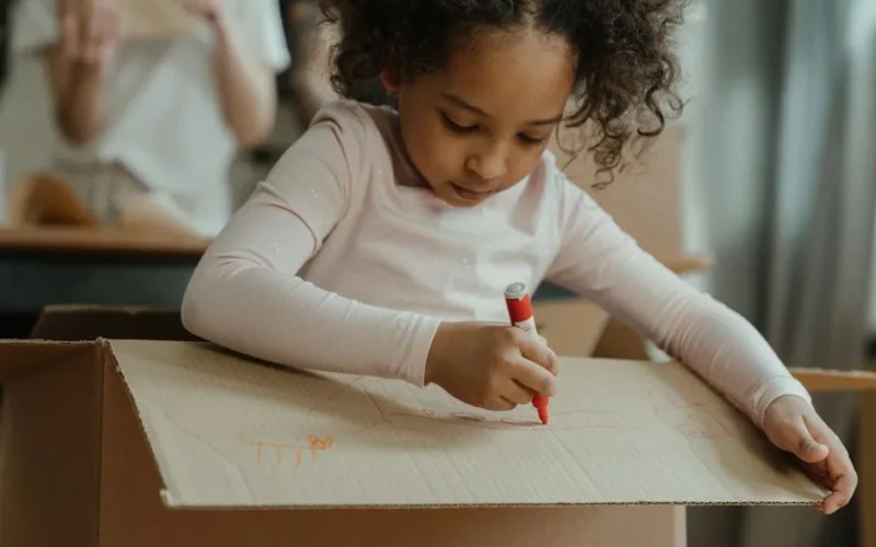 Young girl engrossed in drawing on a cardboard box during a creative indoor activity.