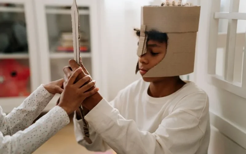 A young child dressed in a cardboard helmet engages in imaginative play with a handmade sword indoors.