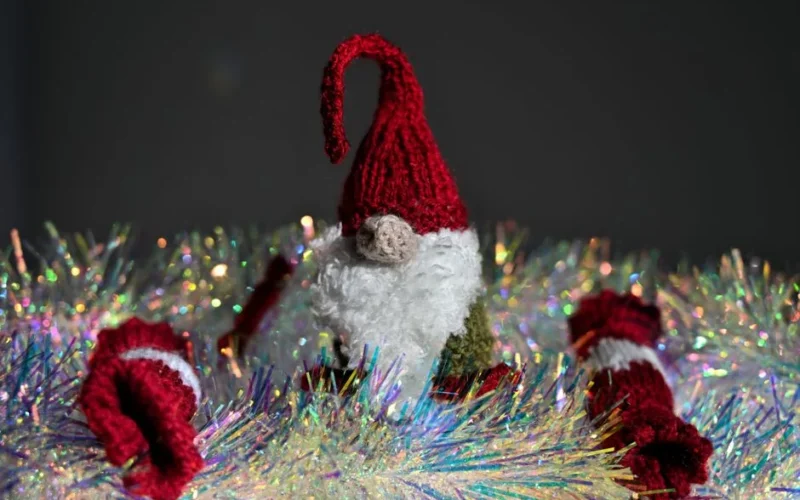 A cute knitted Christmas gnome stands amidst colorful tinsel, creating a festive holiday atmosphere.