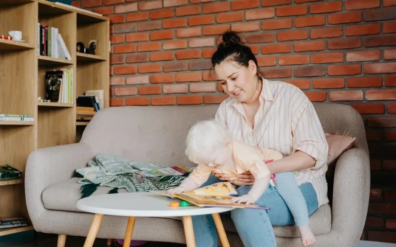 A mother lovingly reads to her baby on a comfortable sofa, surrounded by bookshelves in a cozy living room.