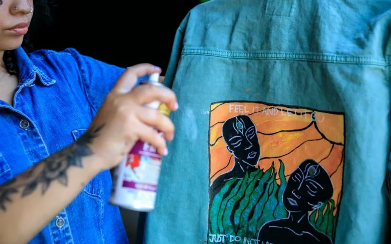 Woman artist spray painting a denim jacket with unique artwork design.