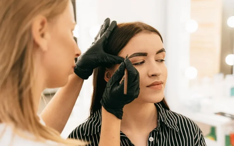 A professional makeup artist shapes a woman's eyebrows in a bright beauty studio.