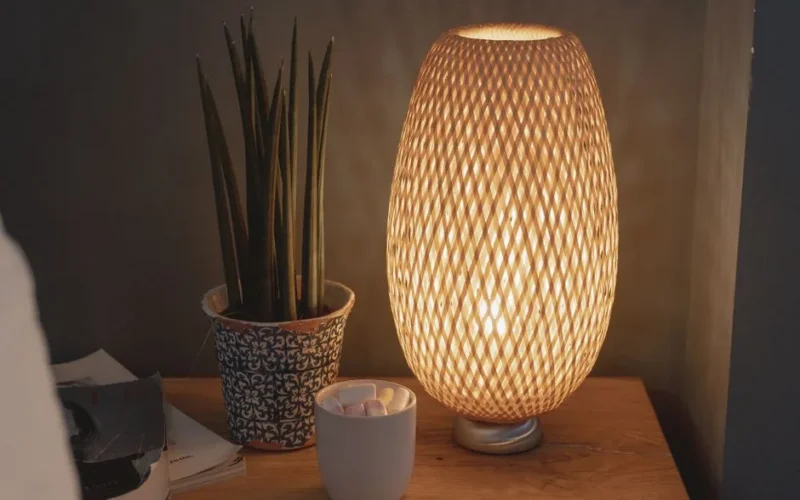 A warm, beautifully lit nightstand featuring a woven lamp and potted plant, creating a cozy atmosphere.