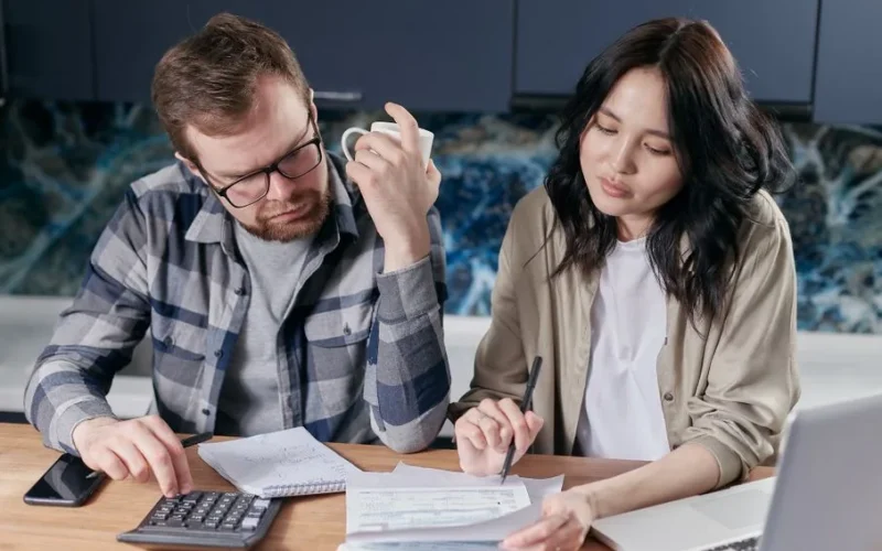 A couple reviewing household bills and budget using a calculator and laptop at their kitchen table.