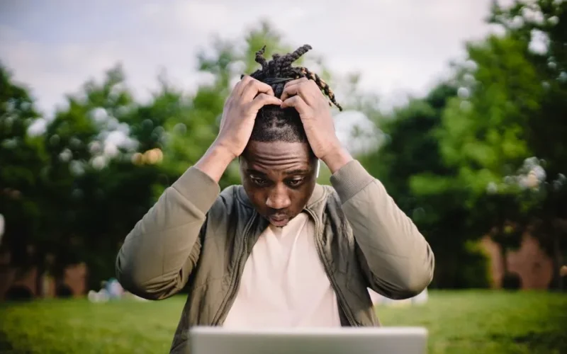 Upset young African American male freelancer clutching head with hands after failure in project while working remotely in park