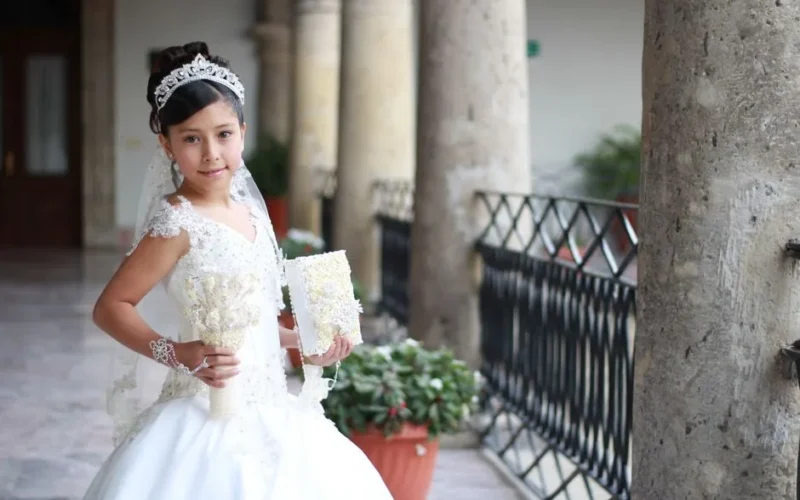 A young girl poses in a traditional white dress for her first communion ceremony, holding a bouquet.