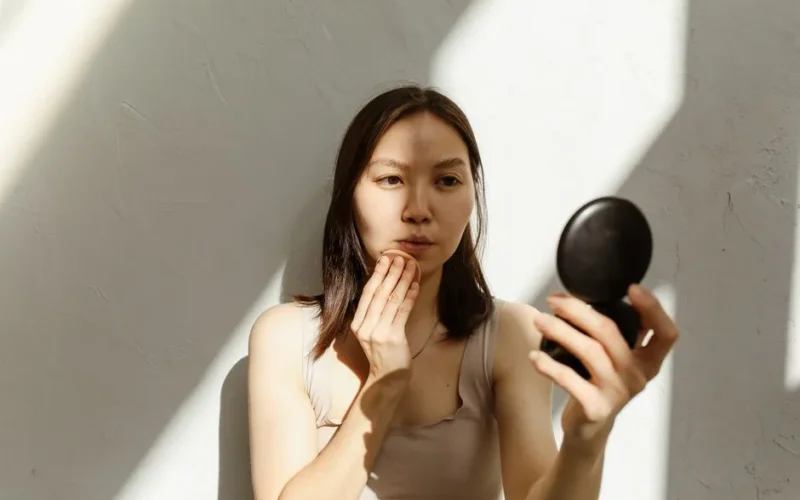 Young Asian woman applying makeup with a mirror in soft natural lighting indoors.