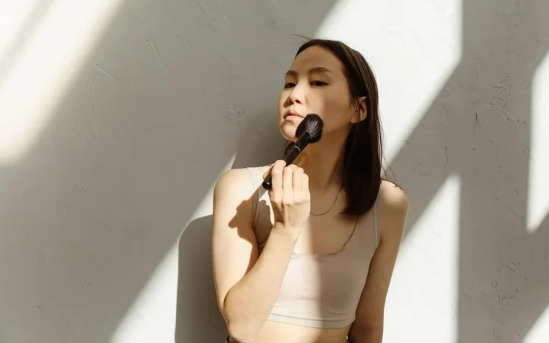Asian woman using makeup brush on face against a sunlit wall indoors.