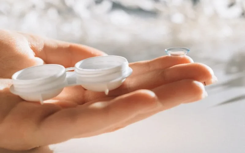 A hand delicately holds a contact lens and case against a blurred background.