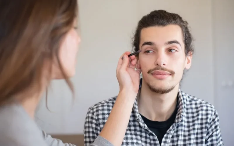 An adult man receives makeup application from a woman in an indoor setting, Portugal.