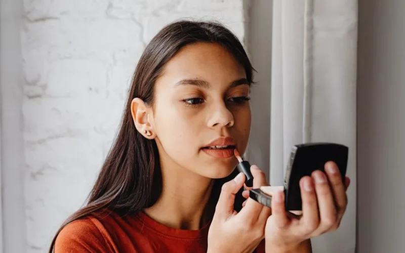 Young woman applying lip tint using a compact mirror indoors by a window.