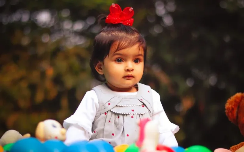 Adorable toddler girl with a red bow, playing with colorful toys in an outdoor setting.