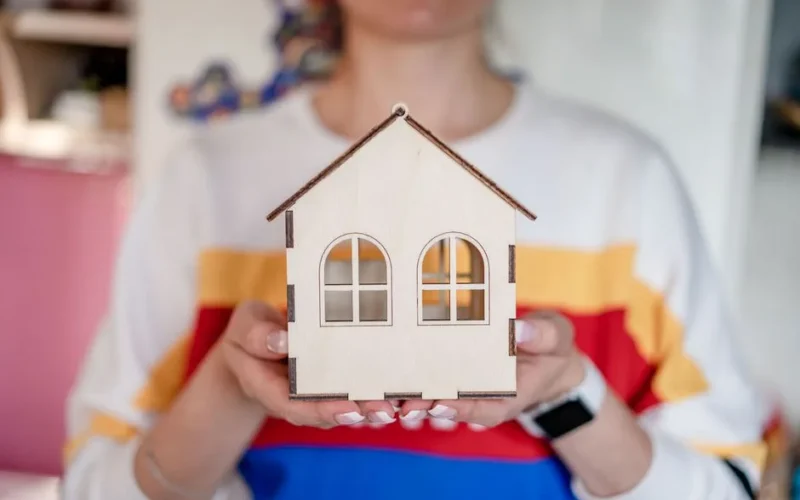 A detailed view of a woman's hands holding a handmade wooden house model indoors.