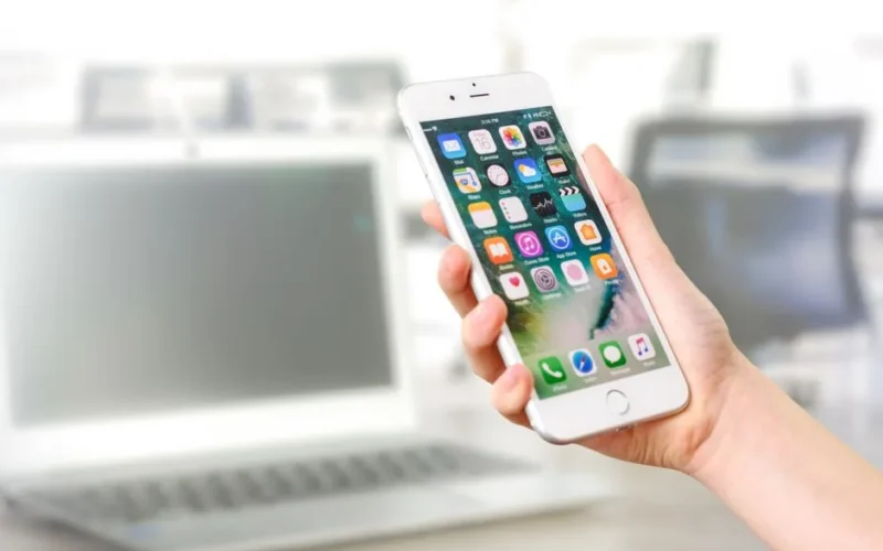 Close-up view of a smartphone displaying apps, held by a hand, with a blurred laptop in the background.