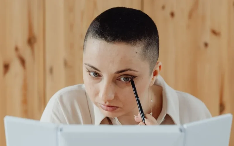 Close-up of woman with short hair applying eyeliner while looking into a mirror.