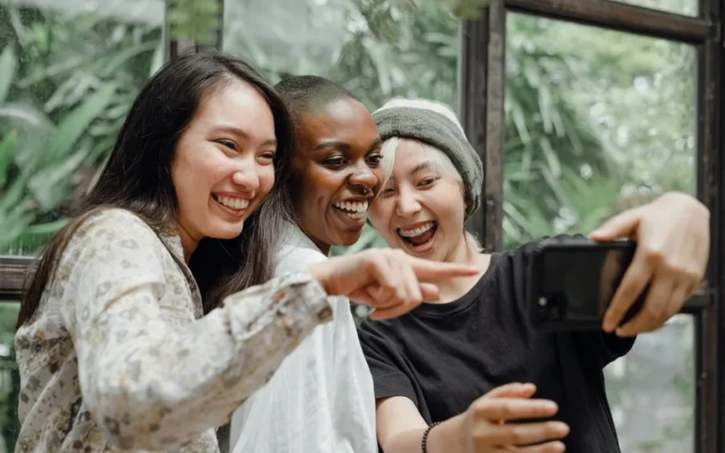 Group of young multiracial females in casual outfits laughing happily at funny selfie just taken on mobile in modern cafe