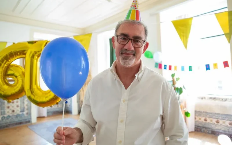 Happy elderly man wearing a party hat holding a blue balloon at a birthday celebration.
