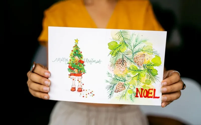 A person holding a Christmas card with festive artwork, including a tree and pinecones.