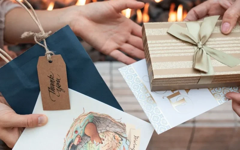 Close-up of hands exchanging beautifully wrapped gifts and thank you cards with a warm fireplace background.