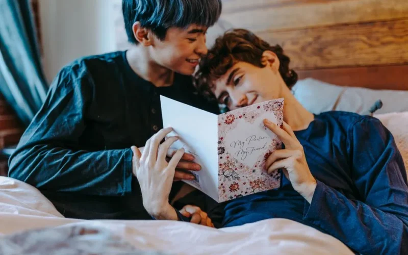 Two young men sharing a tender moment reading a greeting card in bed.