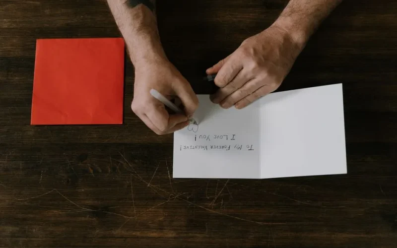 Man writing a romantic message on a card with a red envelope, top view.