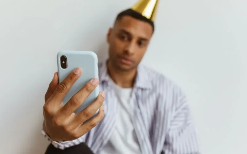 A man wearing a party hat takes a selfie with his smartphone indoors against a plain white background.