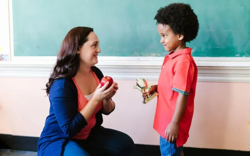 A teacher presents a student with an apple and trophy in a classroom setting.