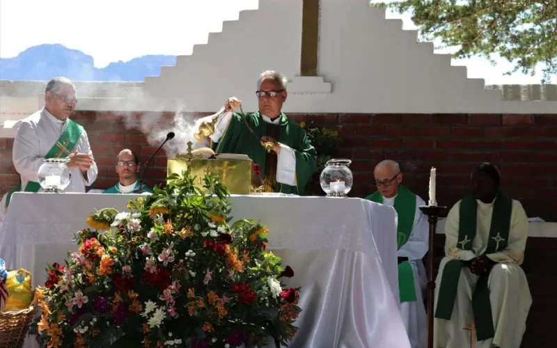 Elderly priest in eyewear and catholic vestment standing with censer near table with Bible and diverse deacons during mass process near colorful bouquet in sunlight in back lit