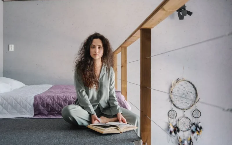 A woman in pajamas sitting on a bed reading a book in a cozy, minimalist bedroom setting.