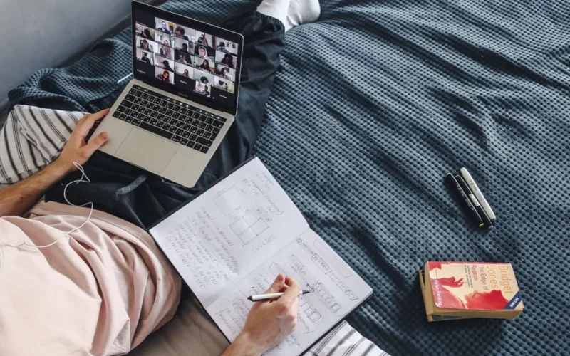 Adult engaged in online class with a laptop and notebook while sitting in a cozy bed setting.