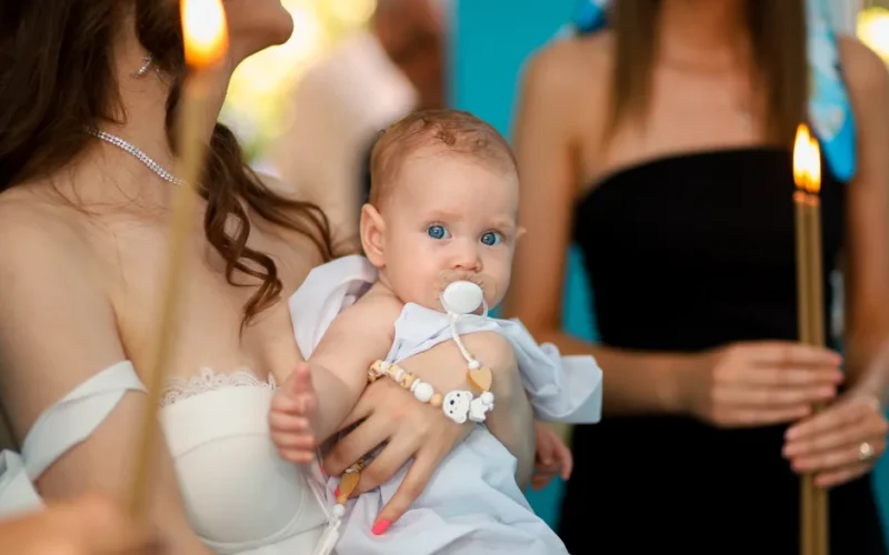 A mother holds her baby during a baptism ceremony with lit candles and warm atmosphere.
