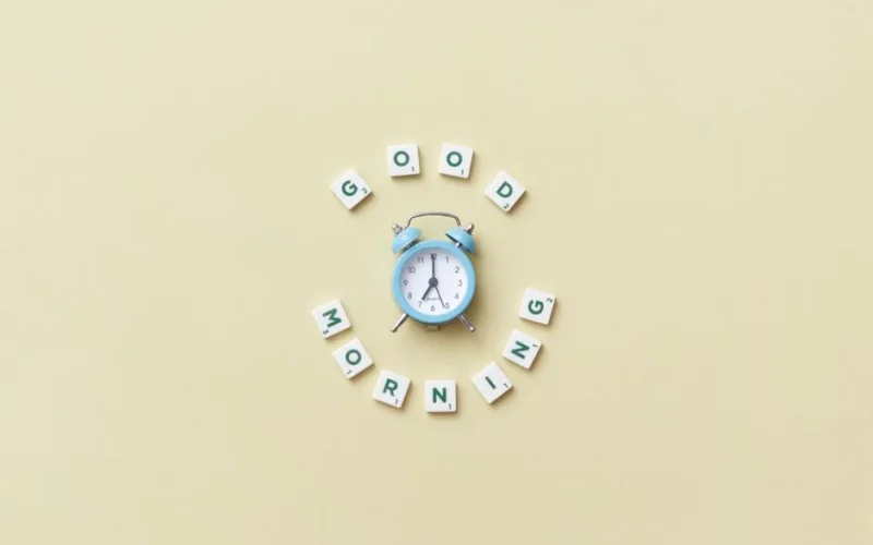 Aesthetic flat lay with a blue alarm clock surrounded by scrabble tiles spelling 'Good Morning' on a pastel background.