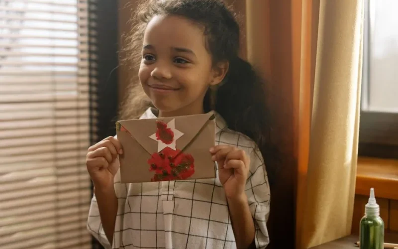 A joyful child smiles while holding a decorated envelope indoors by a window.