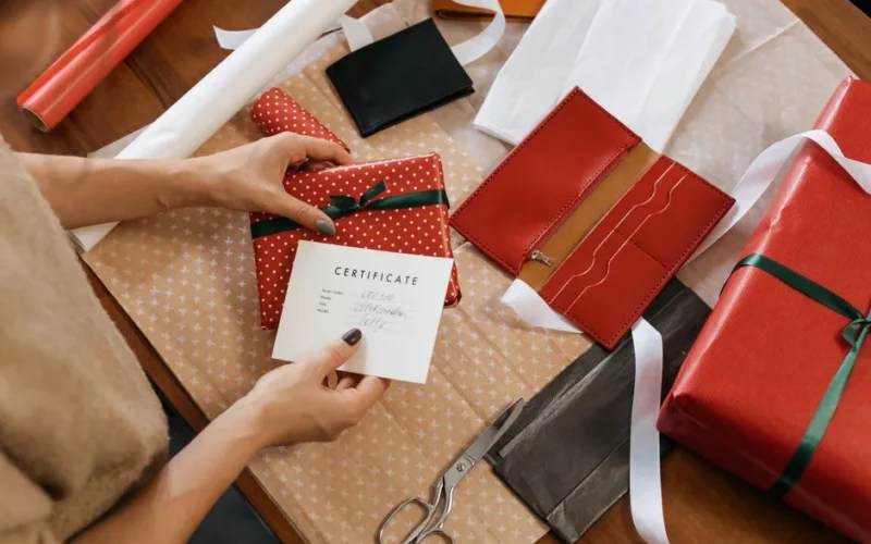 Hands wrapping gifts with a certificate on a wooden table.