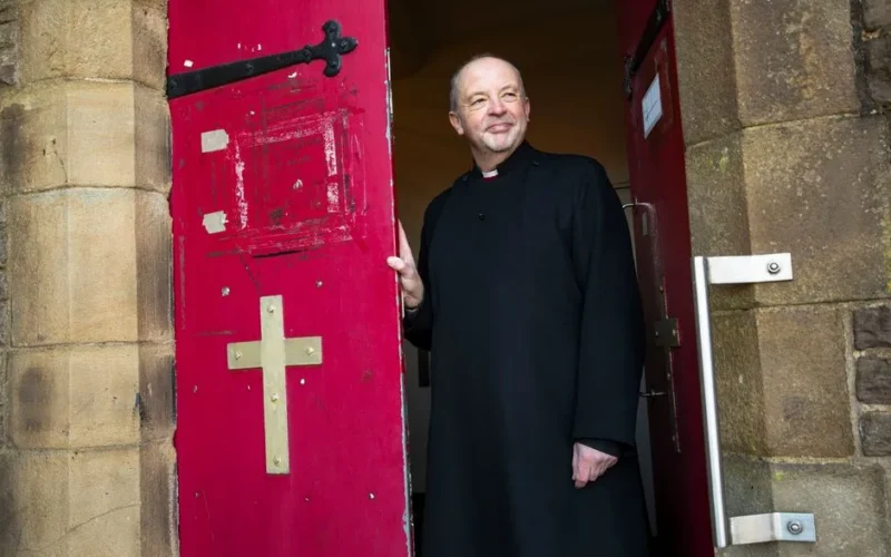 A smiling priest stands in a welcoming pose at the entrance of a church with bright red doors.