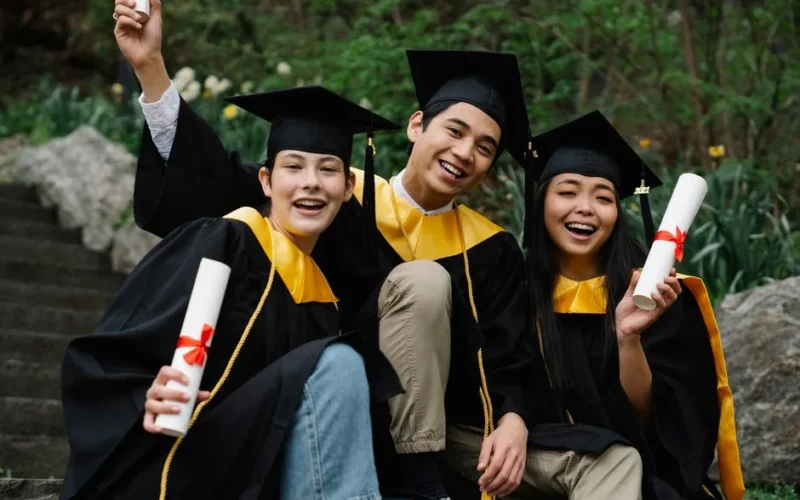 Three diverse graduates joyfully celebrating outdoors in caps, gowns, and holding diplomas.