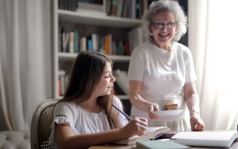 Cheerful grandmother in glasses and casual clothes smiling at camera while giving pastry on plate and mug of tasty beverage to joyful teenager sitting at table with books and exercise book and study in light cozy living room