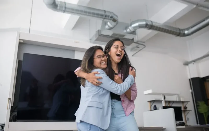 Two women confidently embrace in a bright modern office, expressing happiness and collegiality.