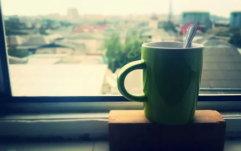 Green mug of coffee by window with urban view, creating a relaxing morning atmosphere.