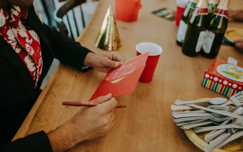 Man writing a Christmas card at a festive holiday party with drinks and decorations.