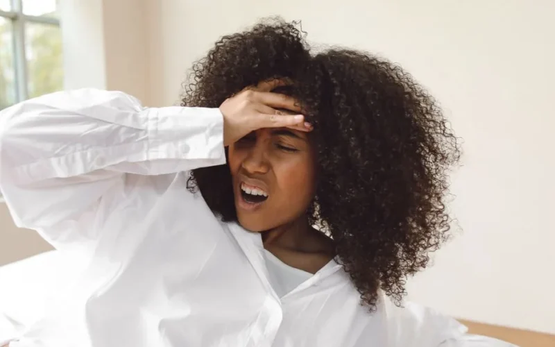 A young woman with curly hair waking up with a headache. She wears a white shirt and seems to be experiencing discomfort.