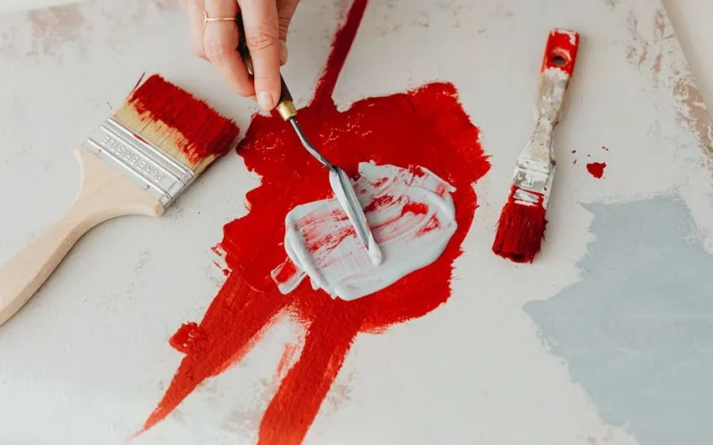 Detailed close-up of hands using a palette knife to mix vibrant red and gray paint on canvas.