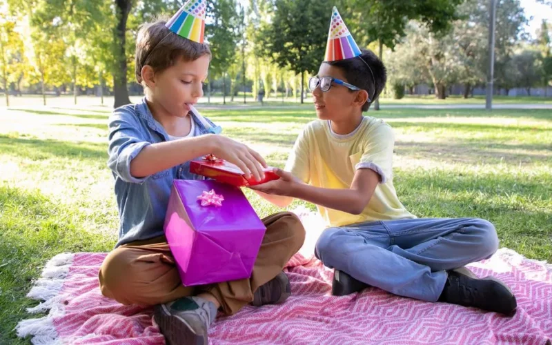 Two children celebrating a birthday outdoors, sharing gifts on a picnic blanket.