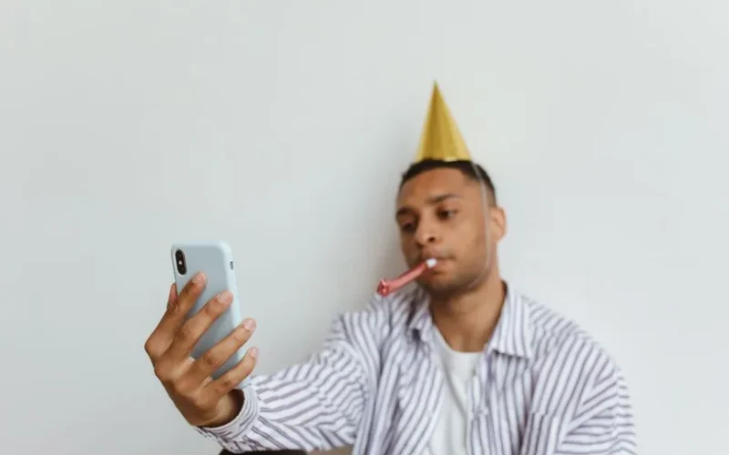 Man with party hat makes video call on smartphone during a virtual birthday celebration.