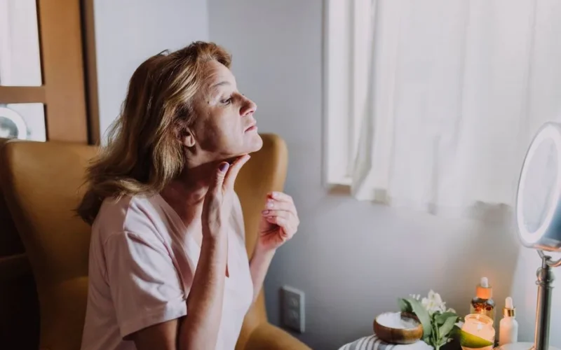 A mature woman examining her skin in a well-lit room with skincare products nearby.